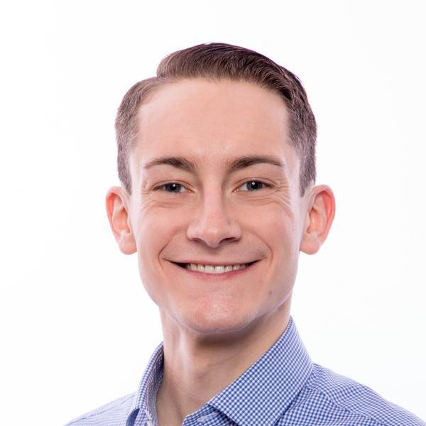 A photograph of Devon King smiling in front of a white background. He is wearing a light blue dress shirt.