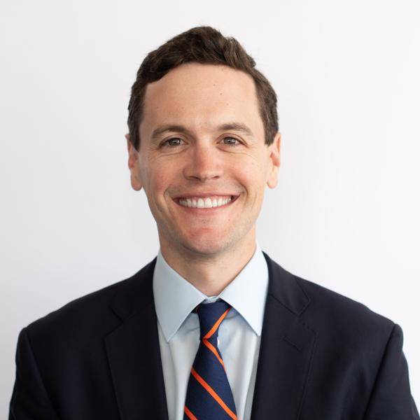 A photograph of Jeff Marr, Jr. smiling in front of a white wall. He is wearing a black suit jacket, a light blue dress shirt, and an orange and blue striped tie. 