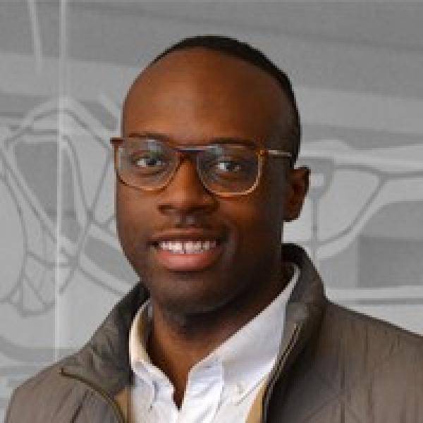 A photograph of Julian Phillips smiling in front of a grey abstractly patterned background. He is wearing glasses, a grey jacket, and a white button-up shirt.