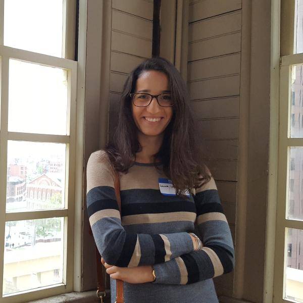 An image of Laura Lacombe smiling to the camera with her arms crossed, standing in a corner of a house.