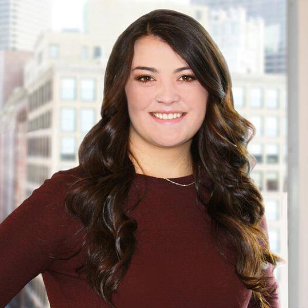 A photograph of Miki Blair standing in front of a window with a city view. She is wearing a maroon shirt.