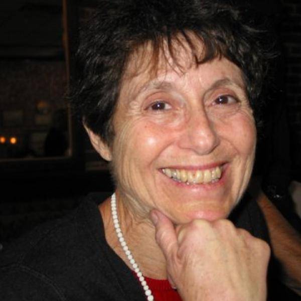 A photograph of Beatrice Nessen smiling. She is wearing a string of pearls, a black sweater, and a red shirt.
