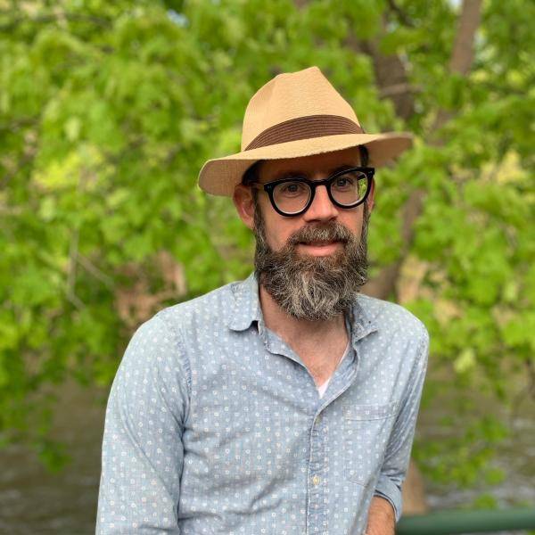 Christopher Scoville leans against a green banister and smiles at the camera. He has a grey beard and a straw hat. He is wearing a denim button-up shirt.