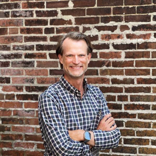 Kevin is smailing at the camera. There is a brick wall behind him.