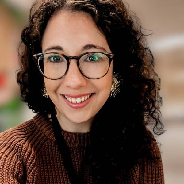 Mallory McCoy is smailing at the camera. She is wearing glasses and a brown sweater. 
