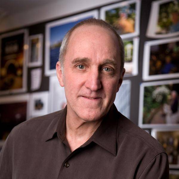 A photograph of Peter Vanderwarker in front of a wall covered in framed pictures. He is wearing a brown long-sleeved collared shirt.
