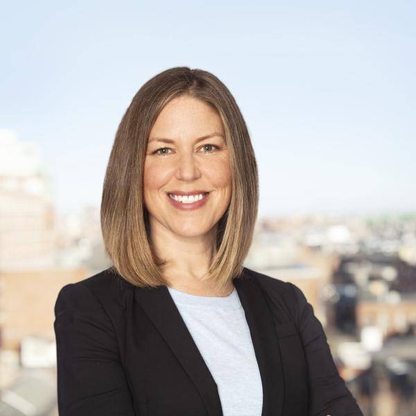 A photo of Regan Shields Ives smiling in front of a cityscape. She has blonde hair and is wearing a black blazer and white shirt.