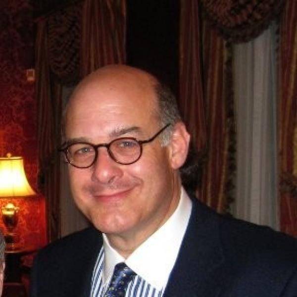 Roger Tackeff smiles for the camera in a dark room. He wears glasses and a suit jacket with a striped shirt and tie.