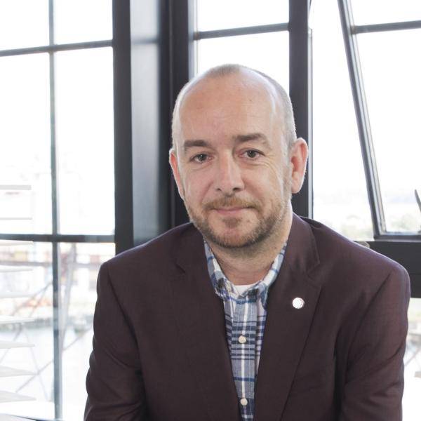 A photograph of Ross Camera leaning on a desk in front of a wall of windows. He is wearing a brown jacket.