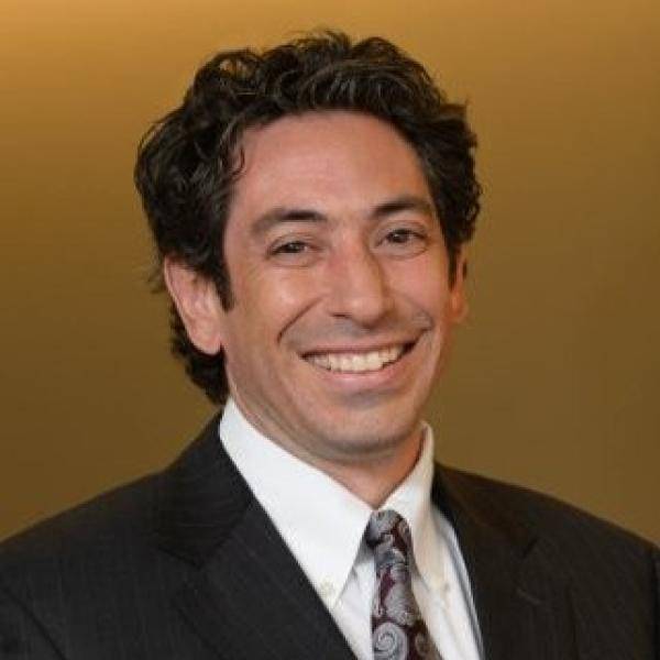 A photograph of Tony Ursillo smiling in front of an orange background. He is wearing a black suit jacket, a white dress shirt, and a grey tie. 