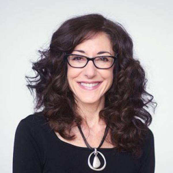 A photograph of Vicki Adjami smiling in front of a white background. She has brown hair and is wearing a black shirt with a large pendant necklace.