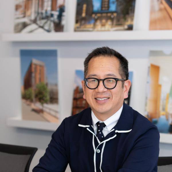 A photograph of Philip Chen smiling in front of a white wall with paintings, he is sitting on a table looking at the camera. He is wearing a blue suit jacket, . He has black hair and glases.