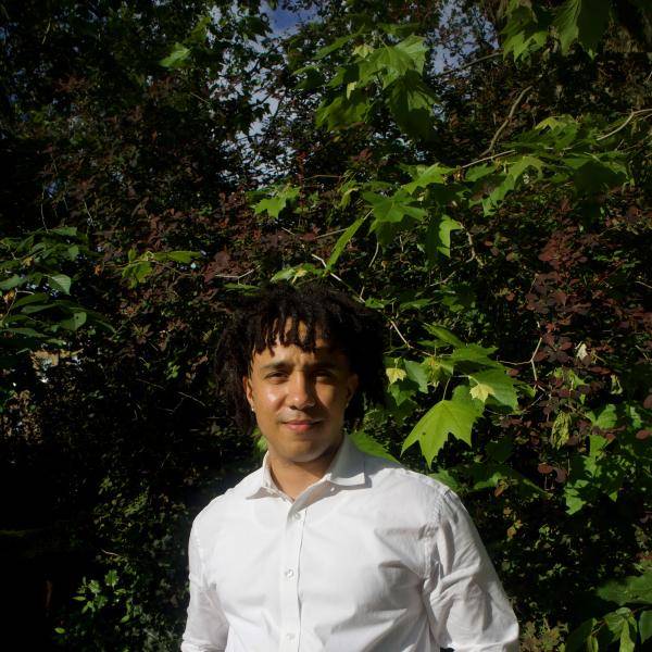 Jon-Lawrence Koloba is looking at the camera in front of nature. He is wearing a white shirt and black pants. He has long and curvy hair.